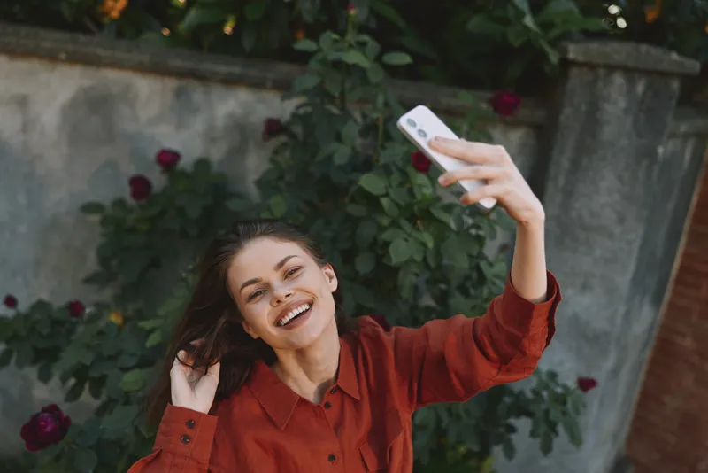 Cheerful woman outdoors, smiling at the camera, taking a selfie in a sunlit moment, red blouse contrasts with lush greenery, relaxed pose, natural colors, carefree mood