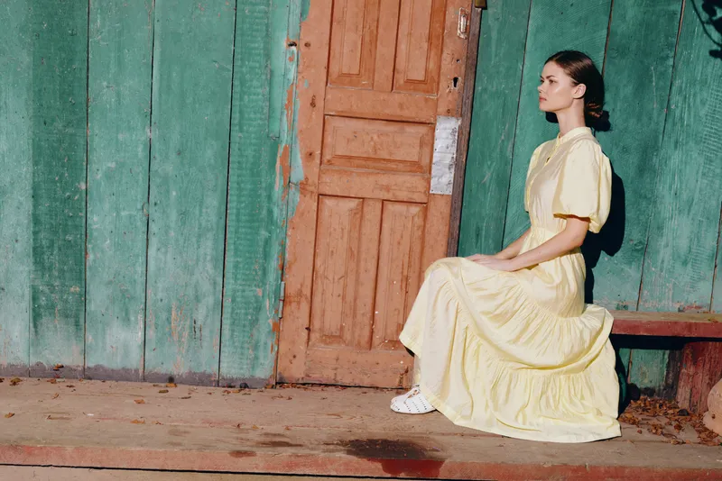 woman sitting on a bench in a flowing yellow dress beside a weathered green wall and wooden door, an outdoor portrait radiating calm, vintage charm, and soft daylight