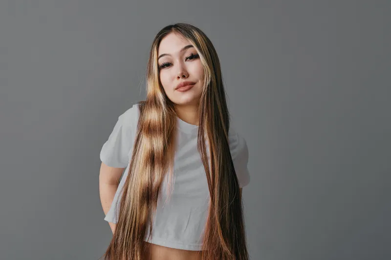 Young woman with long straight hair wearing a white crop top posing against gray background in studio. Female model with confident expression and casual style portrait.