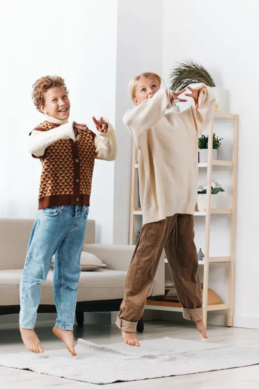 Two young boys jump with joy in a bright living room, wearing cozy sweaters and denim, capturing a playful moment and carefree energy in a warm home setting.