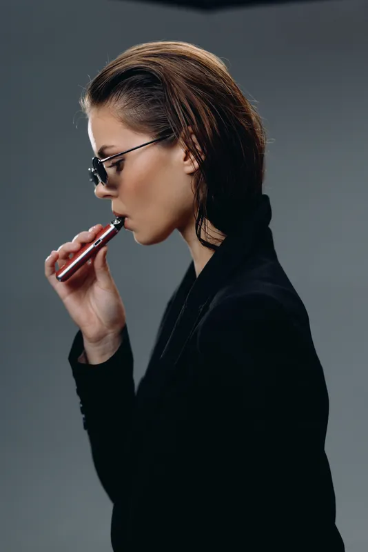 Stylish woman vaping with sunglasses wearing black jacket in studio against grey background
