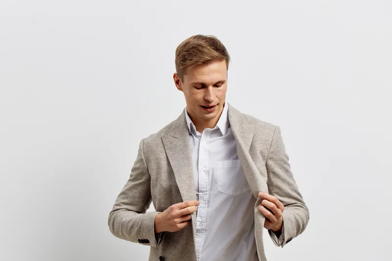 Confident young man adjusting grey textured blazer over white shirt isolated on plain white background studio portrait. Fashion and style concept with professional appearance and modern menswear.