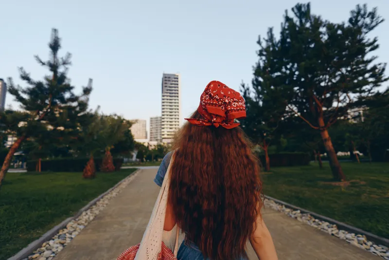 woman walking outdoors with long hair and red headscarf in city park during daytime clear sky