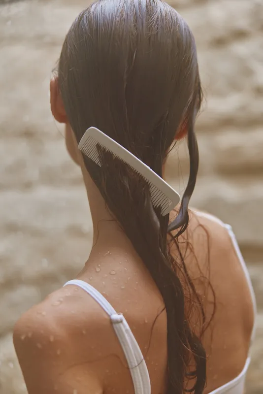 Woman combing wet long hair with white comb, back view of slender female with droplets on skin, natural light, soft neutral background, hair care concept.