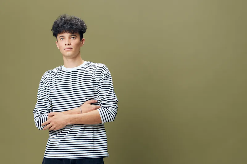 young man with curly hair wearing striped shirt stands with arms crossed on olive green background, casual style and confident expression, studio portrait