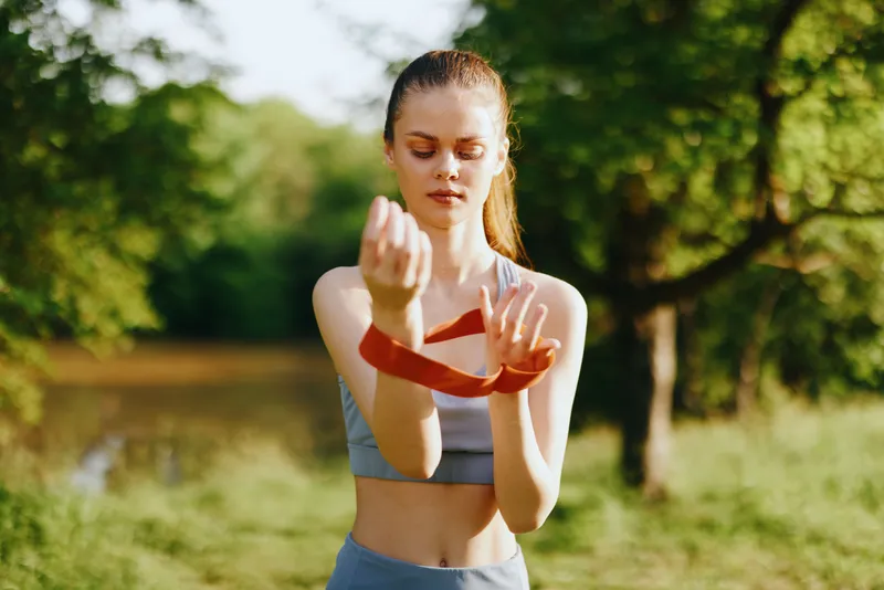 Young woman in activewear applying resistance band outdoors, showcasing fitness and confidence in a natural setting surrounded by greenery