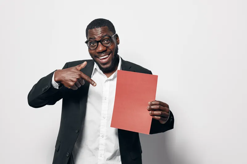 African American man with dark skin wearing glasses and black suit pointing at red paper sheet with smile on white background. Business casual style and confident expression.