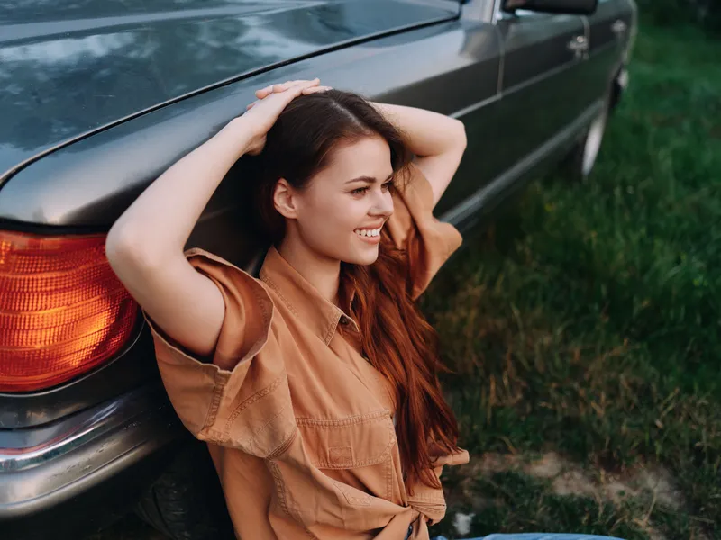 Young woman smiling beside a classic car in a sunny field, wearing a casual brown shirt, showcasing joy and freedom in a serene outdoor setting.