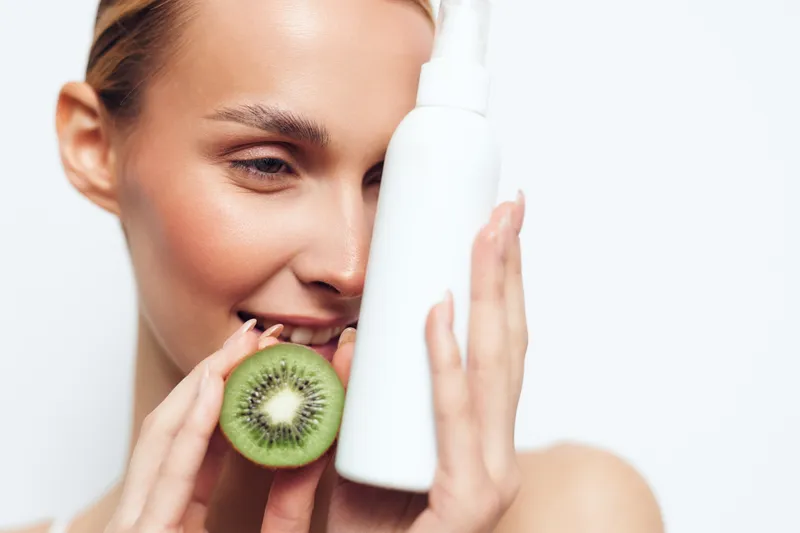 Young woman with natural skin holds white cosmetic bottle and fresh kiwi fruit close to face smiling with closed eyes isolated on white background for skincare concept.