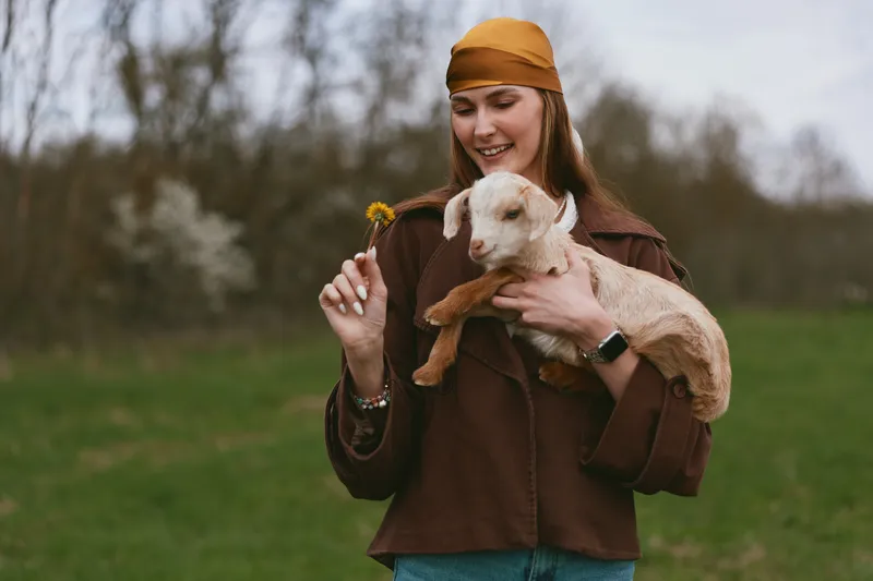 Joyful woman outdoors holding a goat kid, flower in hand, smiling with the animal close, casual jacket and hat, sunlit meadow, warm natural colors, relaxed lifestyle moment.