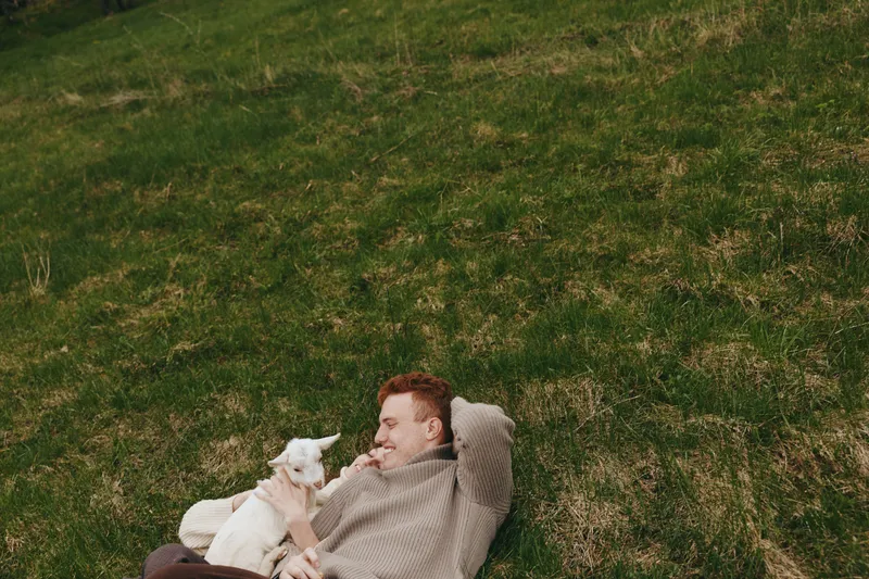 Man outdoors, relaxing on grass, smiling toward a curious goat kid, cozy sweater, gentle breeze, sunlit moment, natural colors, tranquil countryside vibe, candid lifestyle scene