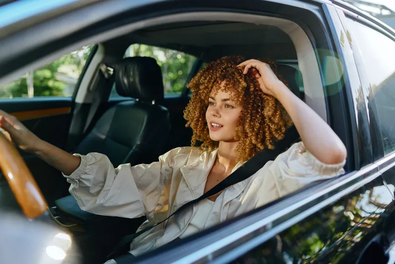 young woman with curly hair driving car during daytime, wearing light blouse and seatbelt, focused on the road with bright natural light and modern interior