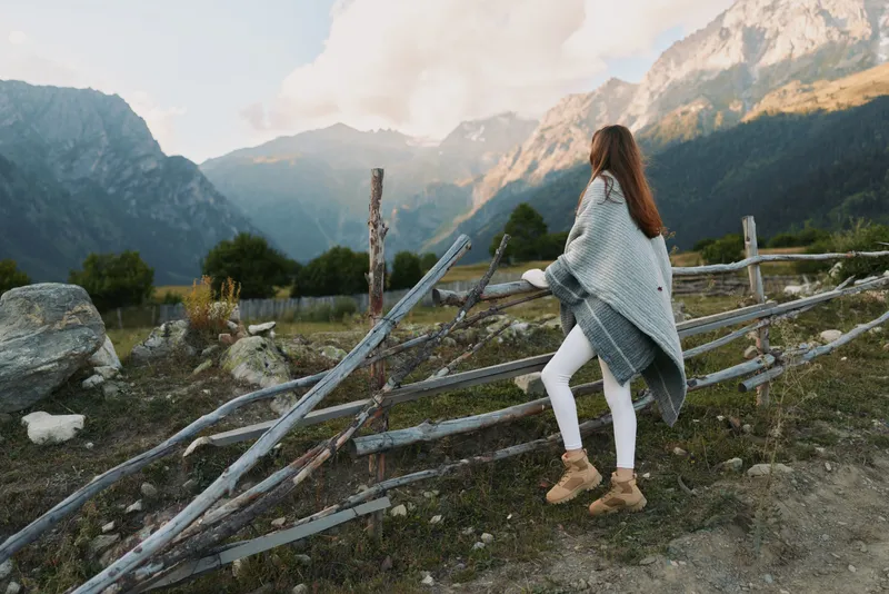 Young woman with long hair wearing a gray shawl and white pants climbs over a wooden fence in a mountainous rural area during daytime.