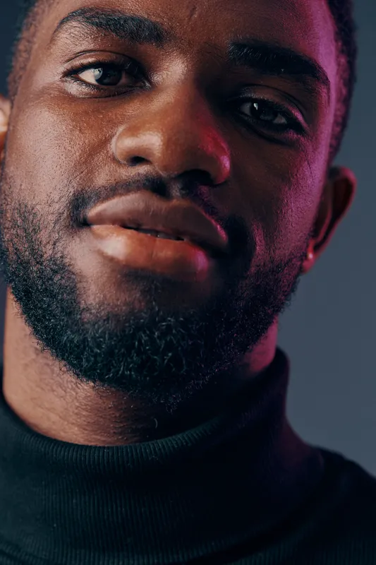 A close up portrait of a confident young Black man in a black turtleneck, showcasing strong facial features and subtle emotions against a softly blurred background