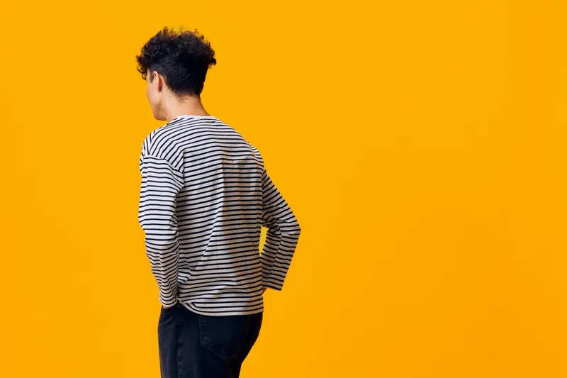 Young man in striped shirt stands with back turned against bright yellow background, casual style, curly hair, minimalist composition, modern and vibrant atmosphere.