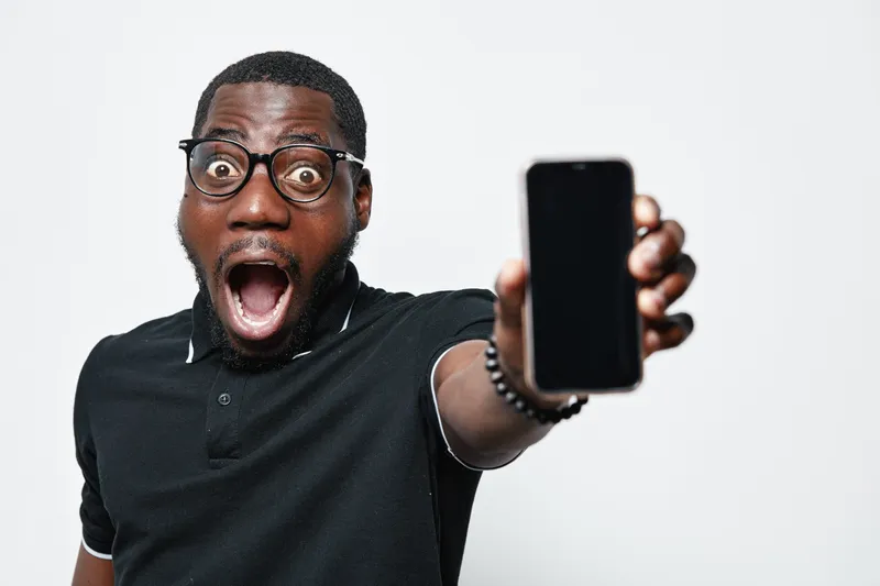 African American man with dark skin and glasses showing smartphone with blank screen, surprised expression and open mouth on white background for technology and communication.