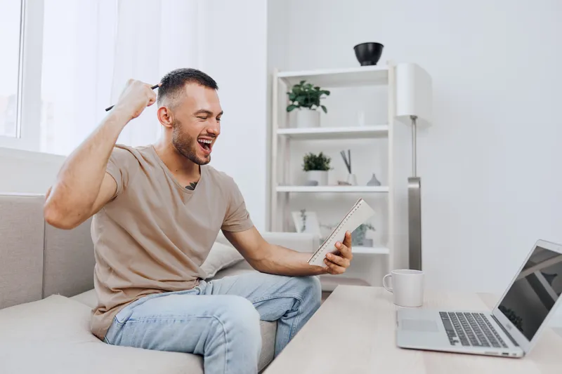 Excited man celebrating success at home, holding a notebook and pen while sitting on a cozy sofa. Bright and modern interior creates an uplifting atmosphere.