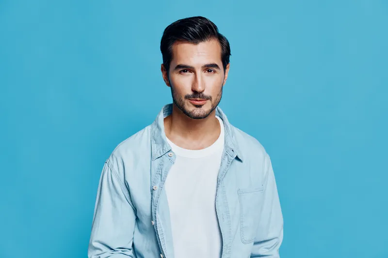 Stylish young smiling man wearing light blue shirt and white t-shirt, posing confidently against a bright blue background in a studio portrait. Fashion concept.