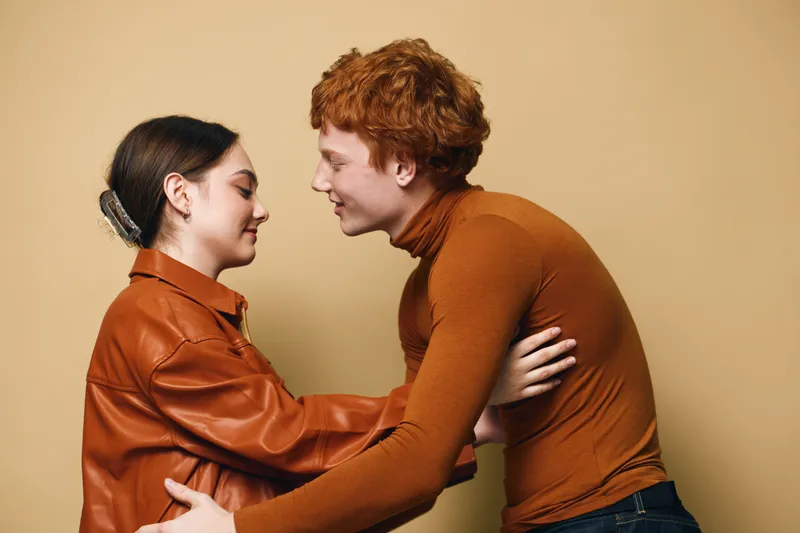 Couple with closed eyes hugging gently in warm brown and orange clothing against beige background. Young lovers sharing peaceful moment with affection, trust and intimacy in casual style.