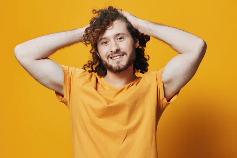 Happy young man with curly hair smiling on a bright yellow background, wearing a casual orange t shirt, conveying positivity and joy