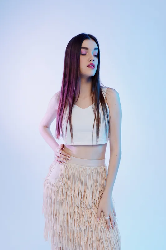 Young woman beauty fashion model with long straight hair poses confidently in a white crop top and fringed skirt on a light background. Studio shot concept.