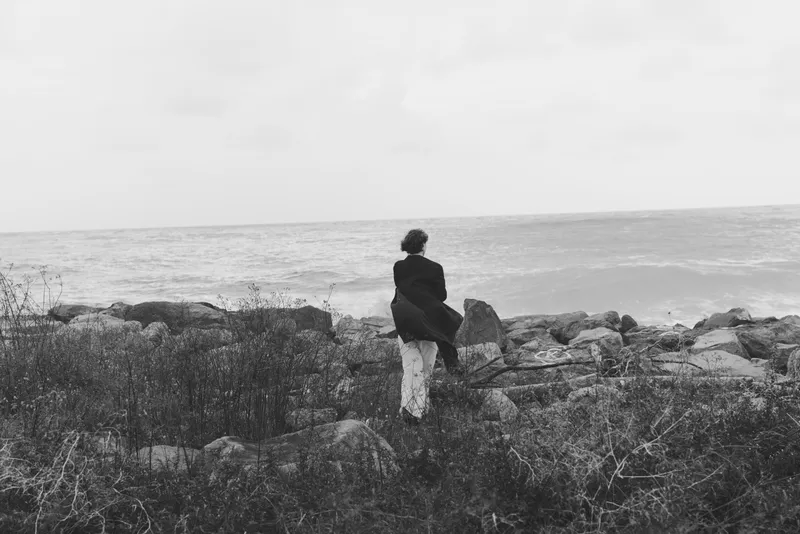Black and white scene of a lone figure standing on rugged coastal rocks, gazing at the sea, wind in the coat, calm horizon and distant waves.