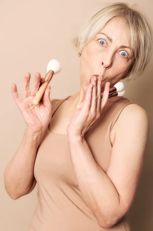 Surprised middle aged woman with short blonde hair holds makeup brushes and covers mouth. Nude background and clothing create soft natural tone. Concept of beauty, skincare, and self care.