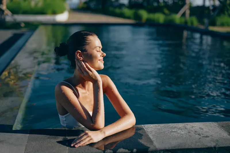 Relaxed woman rests beside a pool, chin in hand, eyes closed, enjoying warm sunlight. Lifestyle moment of wellness, serenity, and outdoor leisure by water.