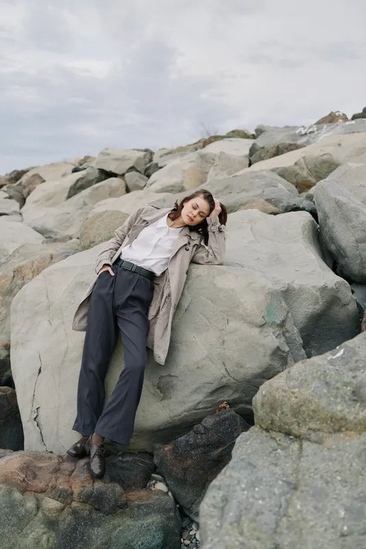 Young woman in casual clothes leans on large rocks outdoors under cloudy sky. Relaxed pose with hand in hair, natural setting with stone textures. Concept of leisure and nature escape.