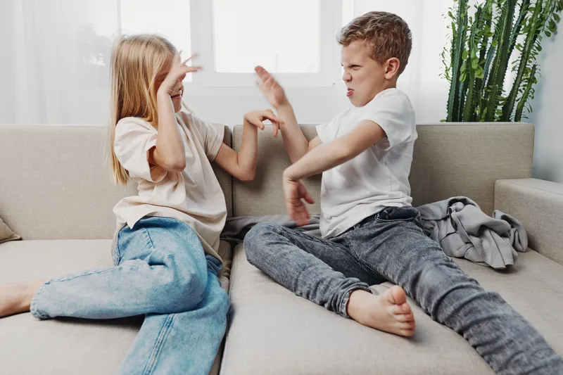 Two children sitting on a sofa playfully pretending to fight with expressive faces and playful gestures in a bright living room setting. Concept of sibling fun and childhood energy.