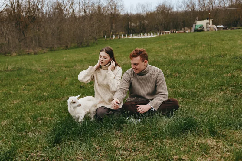 Casual couple sits on a grassy field with a small white dog, wearing knitted sweaters, relaxed mood, peaceful afternoon outdoors in a park, intimate moment of companionship and calm.