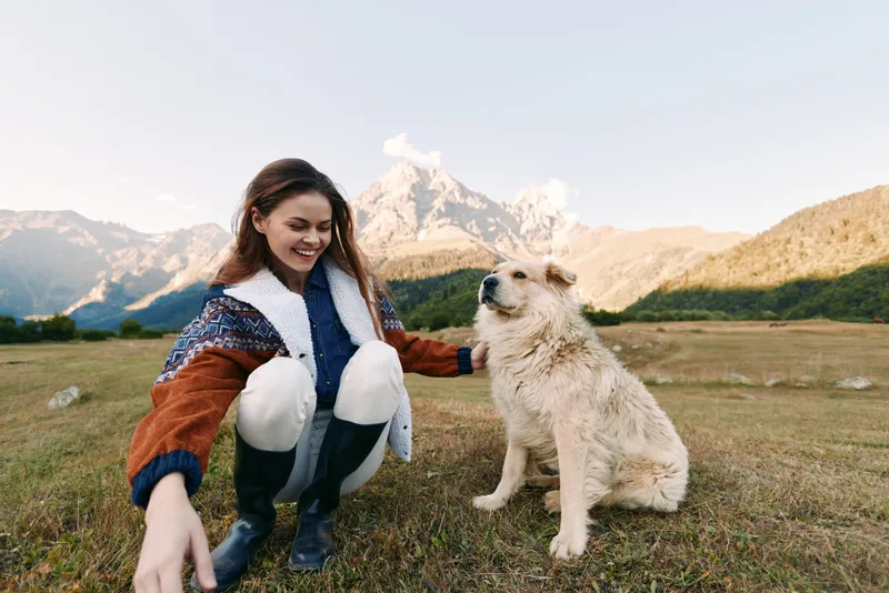 Woman, dog, mountains, meadow, outdoor, pet, happy, nature — Young woman crouching and smiling beside fluffy dog on grassy meadow with mountain backdrop, enjoying hiking and pet companionship.
