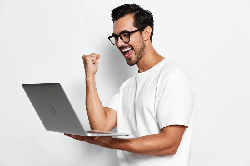 Excited young caucasian man celebrating success while holding a laptop, wearing a casual white t-shirt against a plain light background. Technology lifestyle concept.