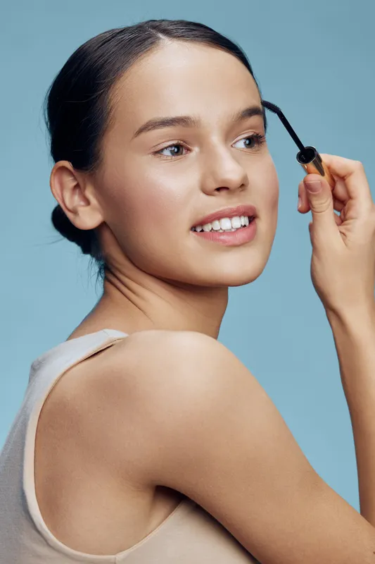 Young woman applying mascara with a smile against a soft blue background, emphasizing beauty and self care in a modern setting