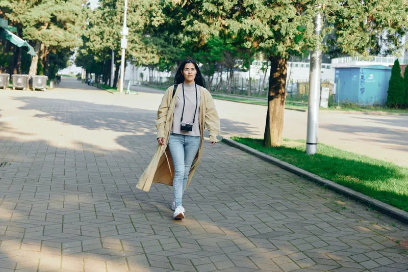 Fashion woman walks along a sunlit street in a long beige coat, casual jeans and sneakers, capturing a relaxed urban moment of style and movement today.