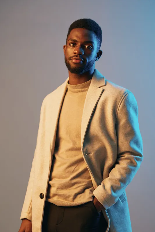 Stylish man in a beige coat and light sweater standing against a gradient background, exuding confidence and modern elegance in a contemporary fashion setting