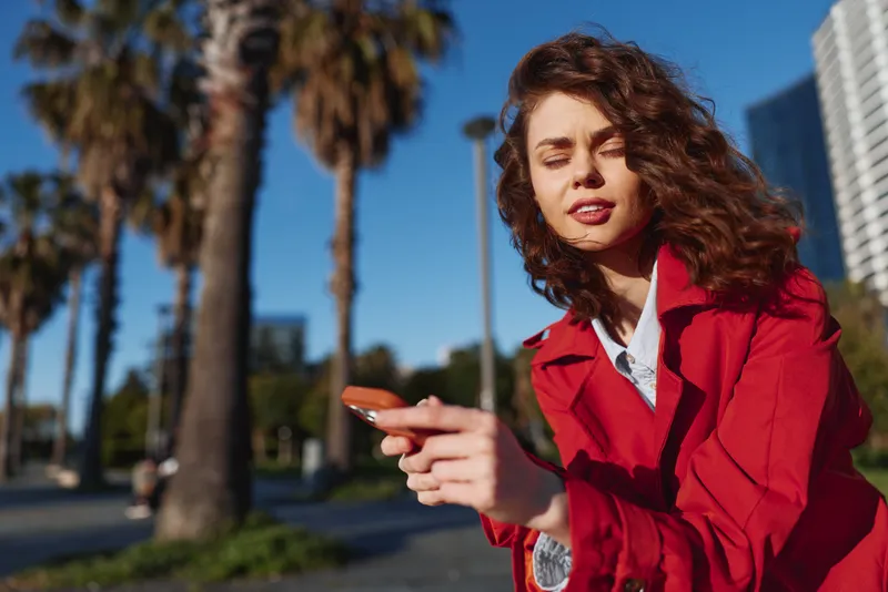 Portrait of a woman with curly hair wearing a red jacket using a smartphone outdoors on a sunny day, palm trees in the background, urban walkway.