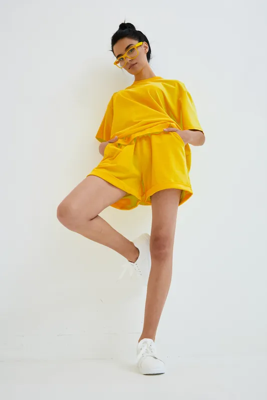 Young woman wearing bright yellow casual outfit and sunglasses poses confidently against white background, showcasing modern minimalist fashion style and relaxed mood.