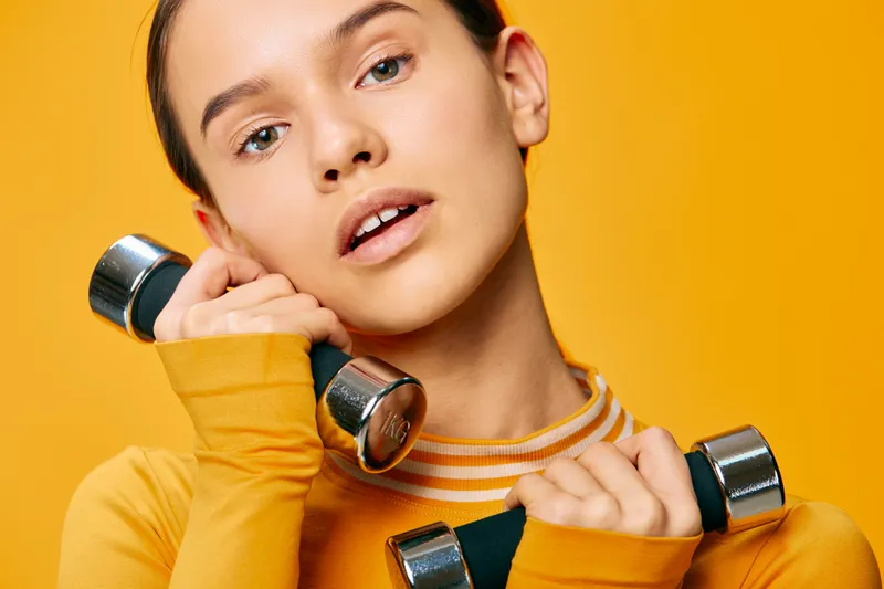 Female fitness enthusiast performing dumbbell workout in a bright yellow studio, focused expression, casual athletic wear, dynamic training moment for health and strength.