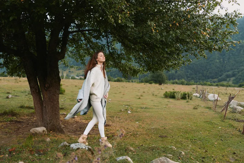 A lone woman in a light coat and white pants strolls beside a leafy tree in a rural meadow, conveying a calm, carefree moment of outdoor lifestyle.