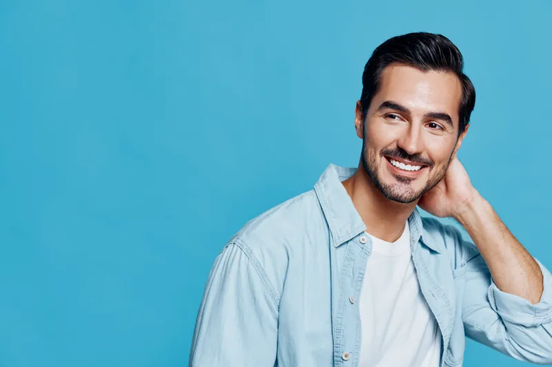 Happy smiling young caucasian man with dark hair wearing light blue shirt, relaxed pose, looking to the side against a solid blue background. People lifestyle concept.