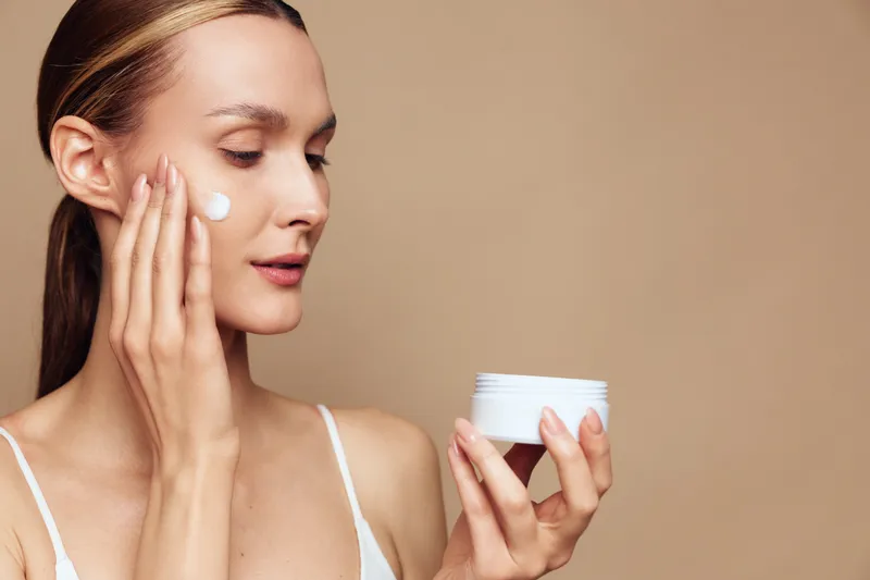 Young woman applying face cream with fingers, skincare routine, natural beauty, soft lighting, neutral background, focused expression, healthy skin care, close-up portrait