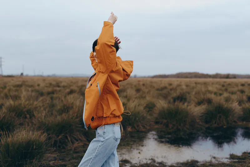 Woman outdoors lifestyle moment, orange jacket and jeans, standing in a grassy field beside a narrow path, cloudy sky, gentle wind, candid relaxed posture, natural colors