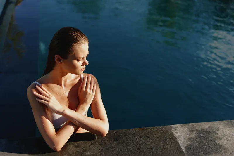 Woman by a poolside, relaxed and thoughtful, sunlit air, warm light, blue water, calm outdoor scene, casual lifestyle moment with soft shadows and calm water, serene vibe