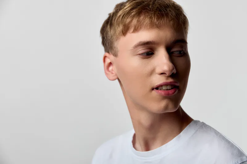 Male, young headshot with face and expression of contemplation, wearing white t shirt, side view against a neutral studio background, subtle mood and clean lighting