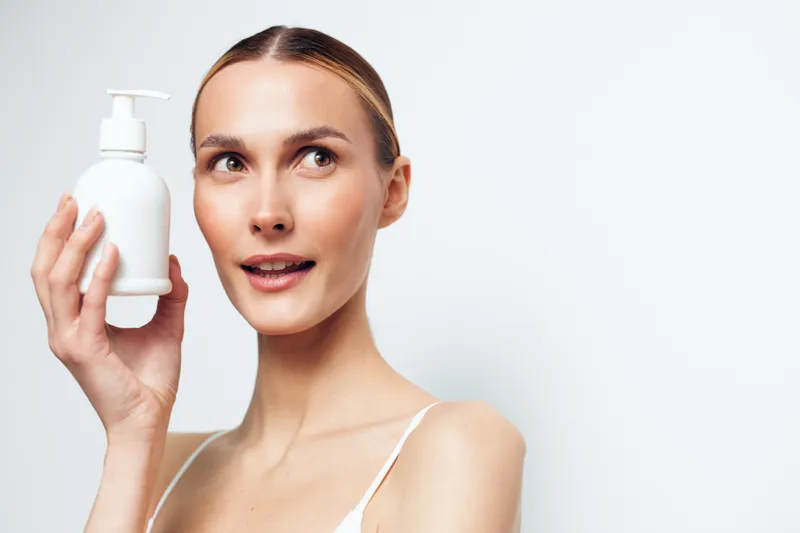 Young woman with clear skin holding white cosmetic bottle, natural makeup, light background, skincare concept, beauty routine, healthy lifestyle, fresh appearance