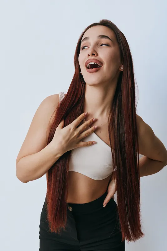 Young woman with long straight hair wearing white crop top and black pants posing with hand on chest and looking up with joyful expression on plain background.