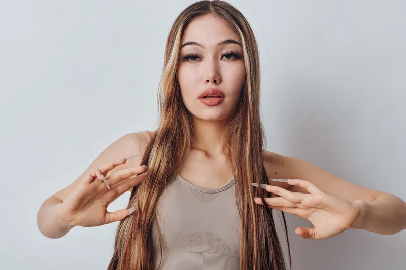 Young woman with long straight hair posing with hands raised in neutral expression studio background. Female model and beauty portrait with natural makeup and casual sleeveless top.