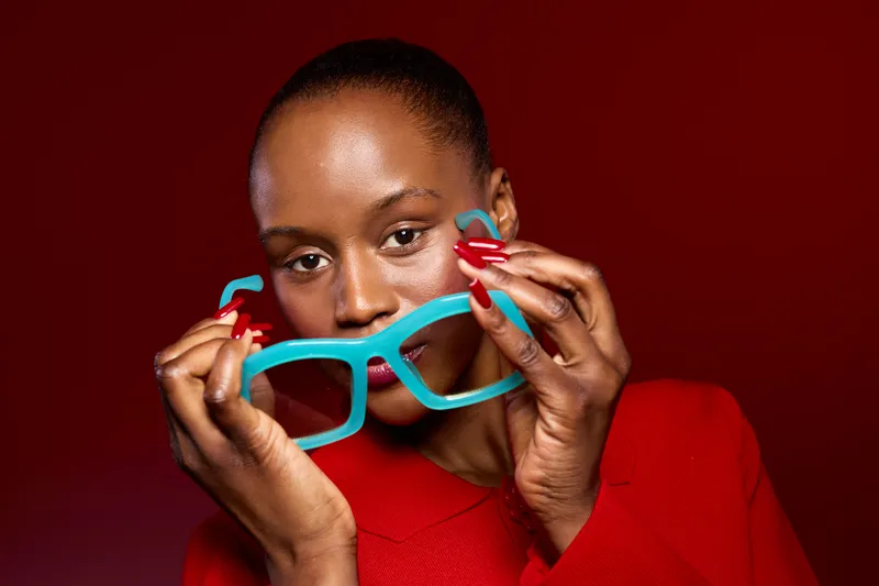 Close-up of a dark skinned African woman holding bright blue glasses in front of her face against a red background. Concept of fashion, beauty, style, and confident female model portrait.