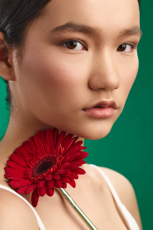Asian woman poses in a studio portrait with a vivid green backdrop, a red flower near her shoulder, soft lighting, calm expression, stylish, elegant, and contemporary style suitable for fashion or